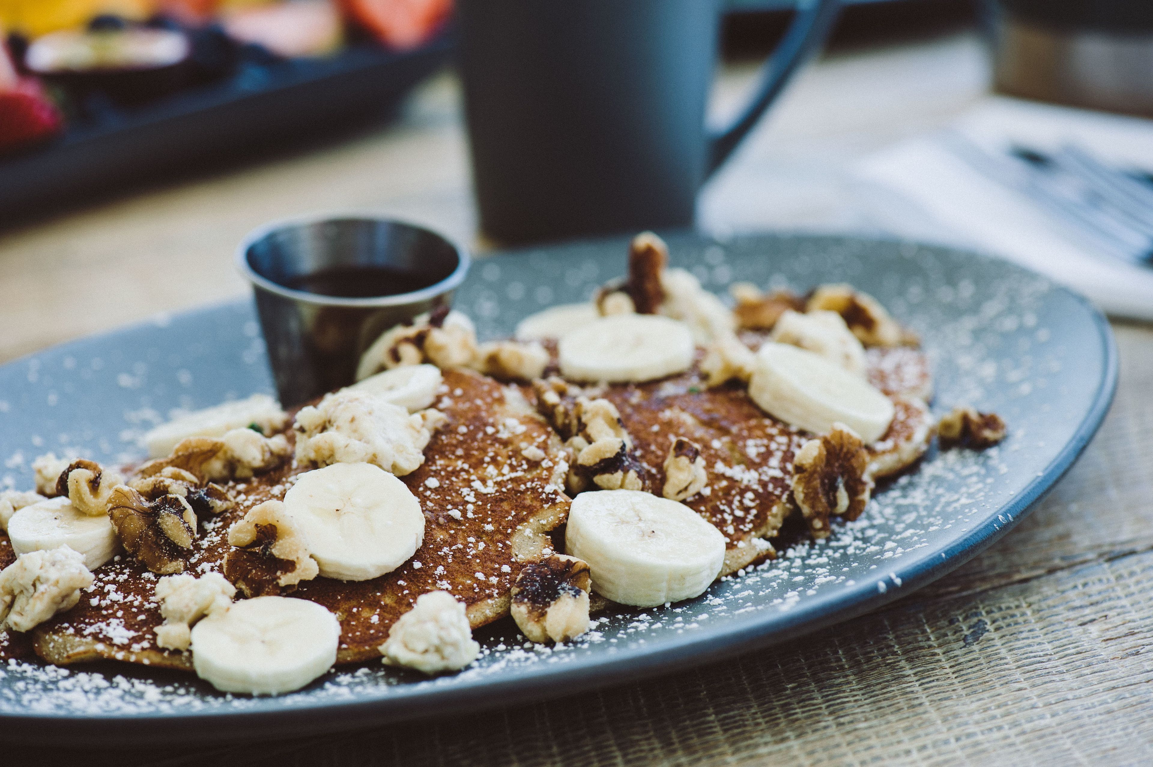 Un plato de tortitas cubiertas de plátanos y nueces, acompañado de azúcar glas.