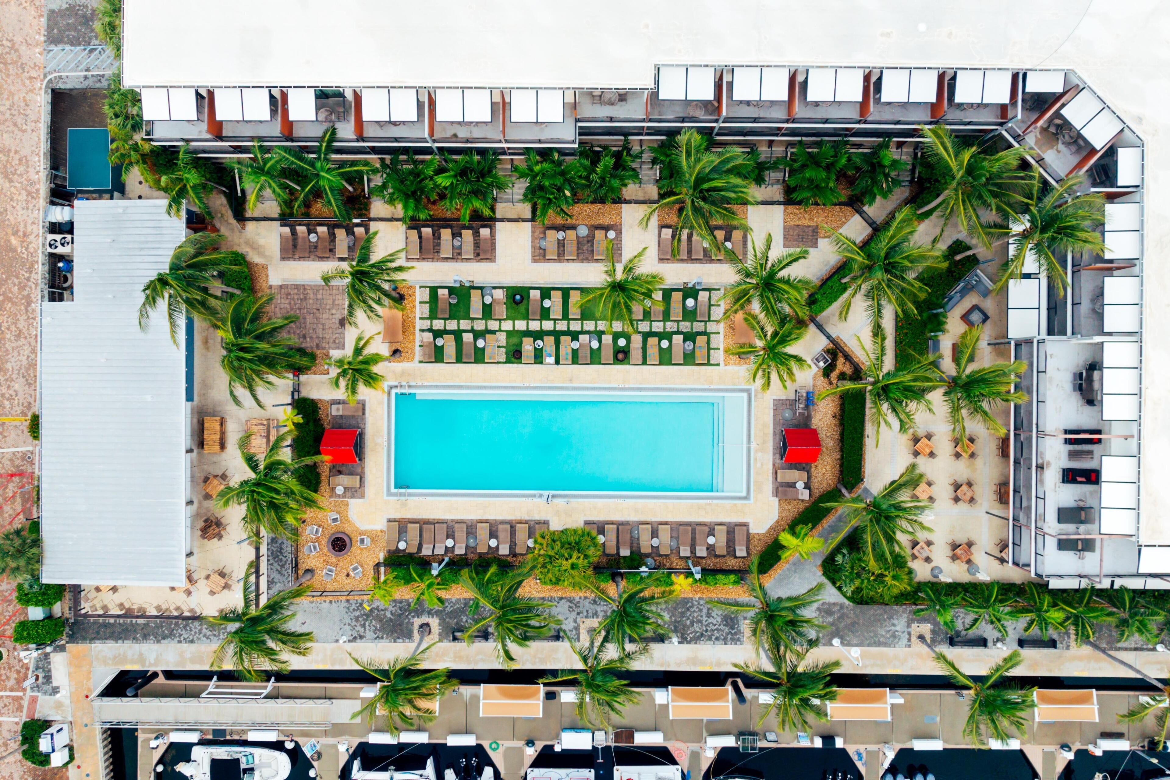 An aerial view of a resort with a central swimming pool, multiple lounge areas, and palm trees.