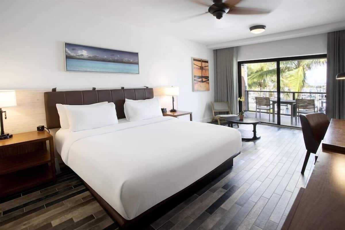 A hotel room with a large bed, a desk, a chair, a balcony with a view, and a ceiling fan.