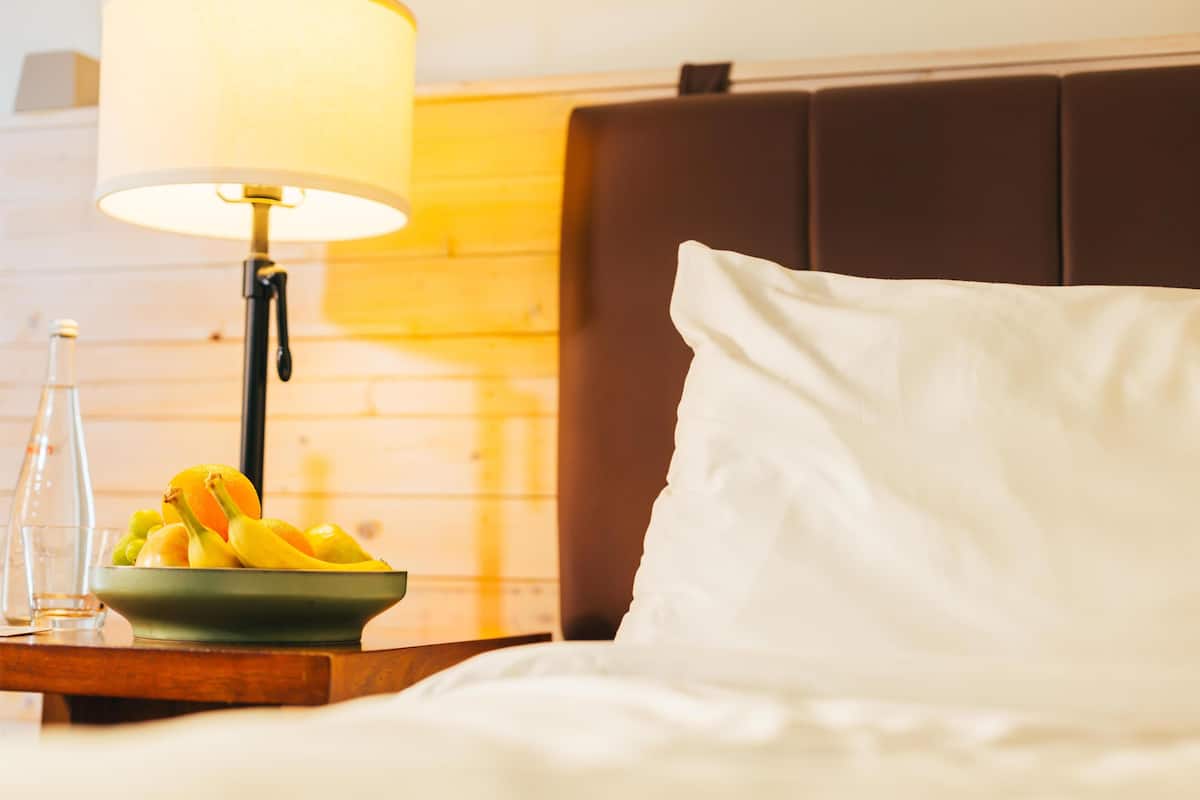 A hotel room with a bed, a bedside table with a lamp, and a bowl of fruit.