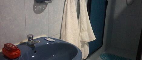 A bathroom with a blue sink, a red soap dispenser, and a glass of water on the sink. There is a towel hanging on the wall and a shower area visible in the background.