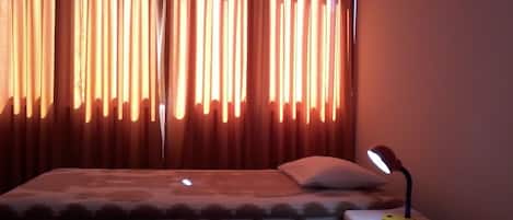 A hotel room with a bed, a bedside table with a lamp, and a window with vertical blinds.