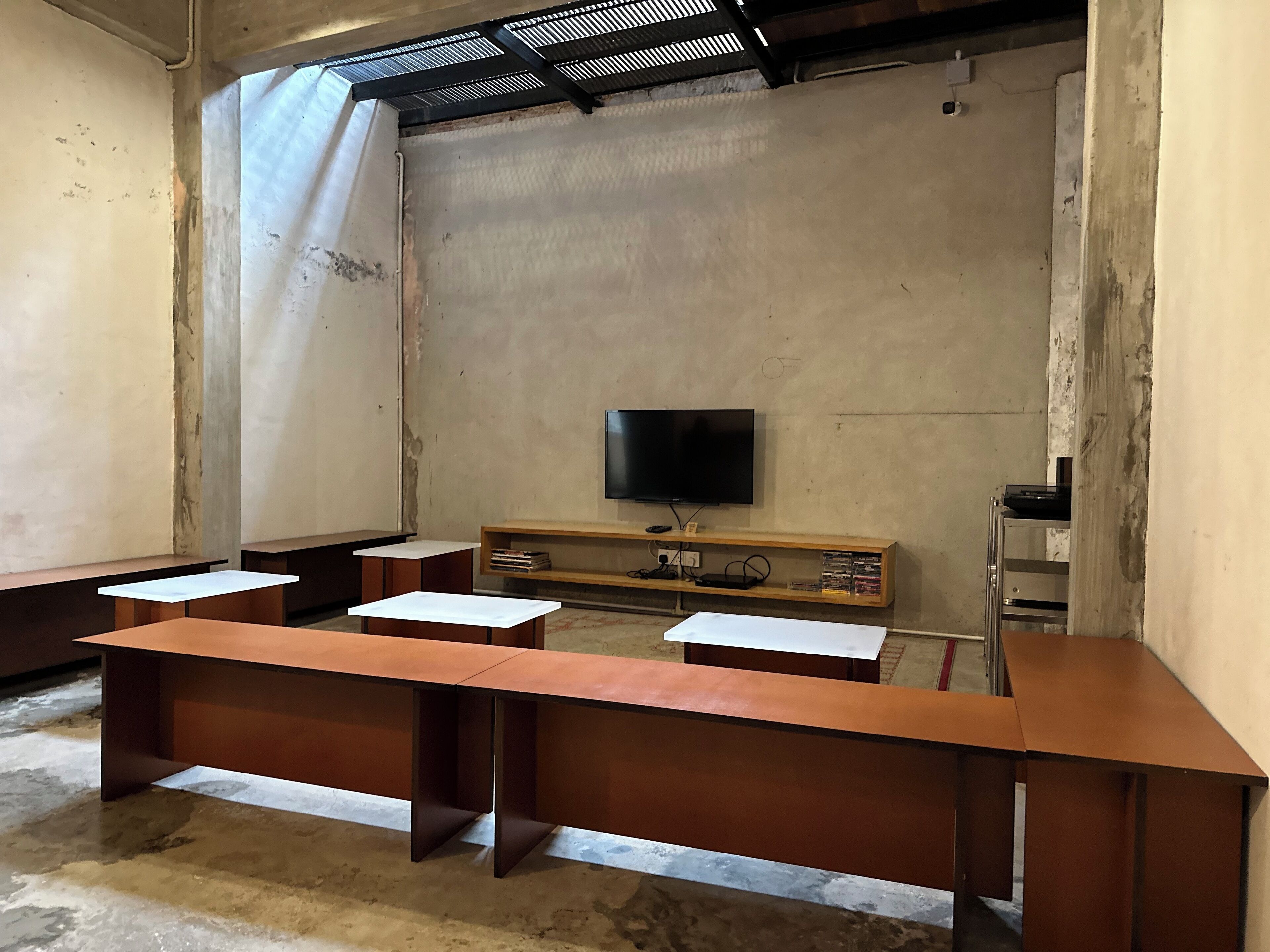 A room with a large flat-screen TV mounted on the wall, wooden desks with white tops, and a wooden cabinet below the TV.