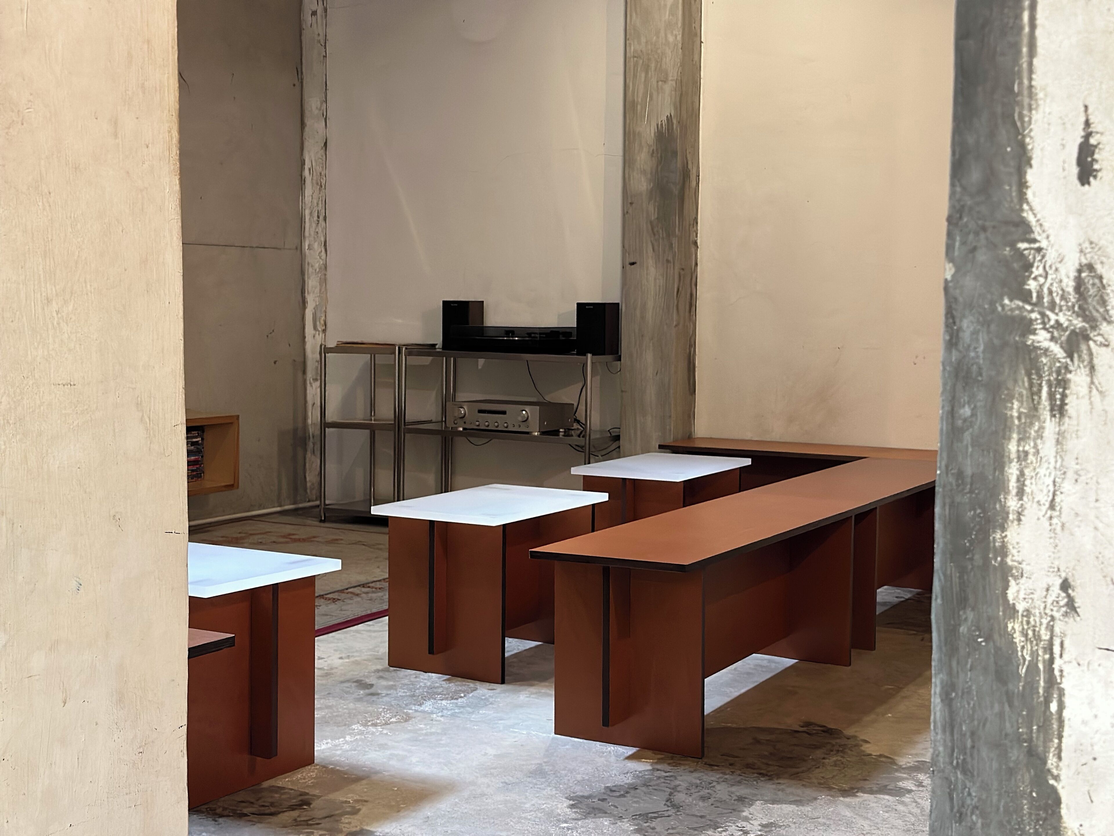 Ένας χώρος με μεγάλα ξύλινα γραφεία και λευκές επιφάνειες, μια μεταλλική βιβλιοθήκη με ηλεκτρονικό εξοπλισμό και ένα τσιμεντένιο δάπεδο.