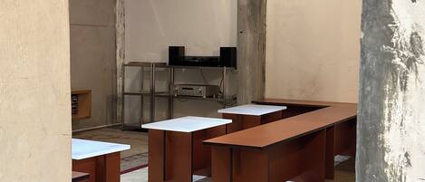 A room with long wooden desks and white tops, a metal shelving unit with electronic equipment, and a concrete floor.