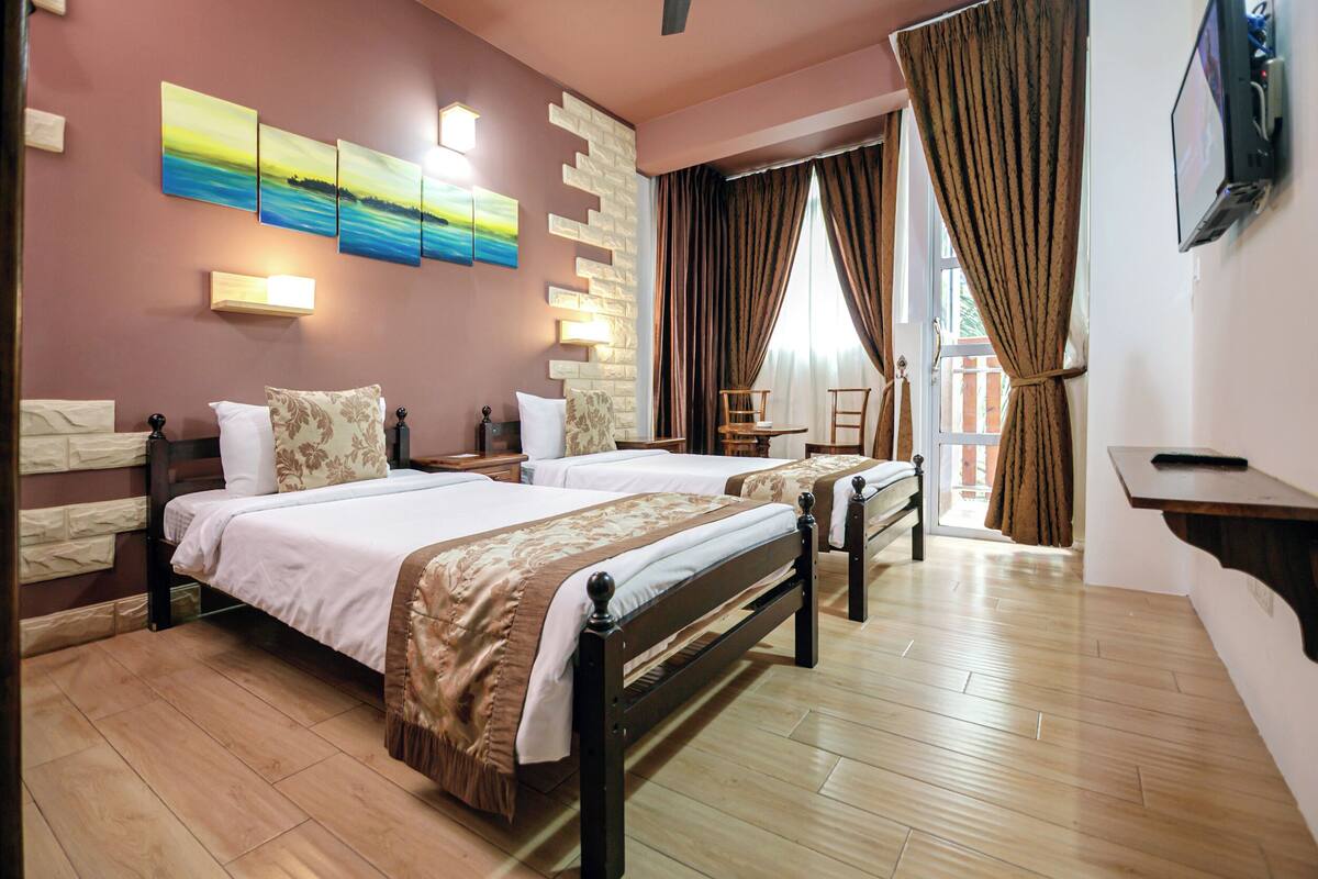 Habitación de hotel con dos camas, piso de madera, televisor montado en la pared y una ventana con cortinas.