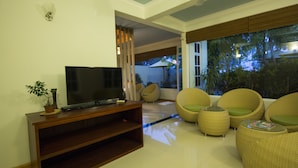 Una sala de estar con un televisor de pantalla plana sobre un soporte de madera, sillas de mimbre con cojines verdes, y un ventanal con vistas a árboles.