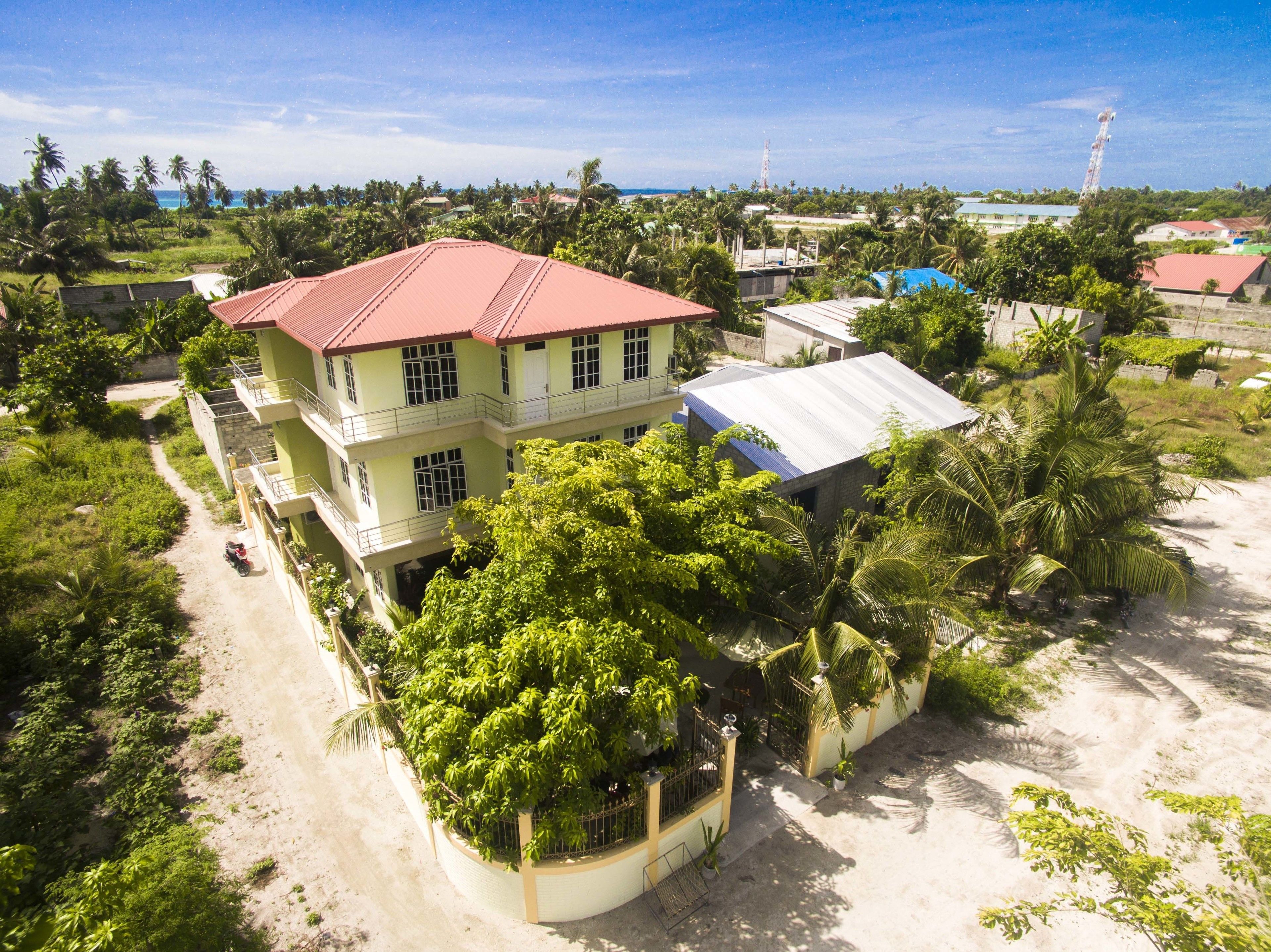 A multi-story building with a red roof, surrounded by greenery and a sandy area.