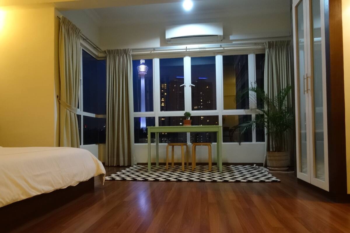A hotel room with a bed, a dining table with two chairs, a potted plant, and a view of a cityscape at night.