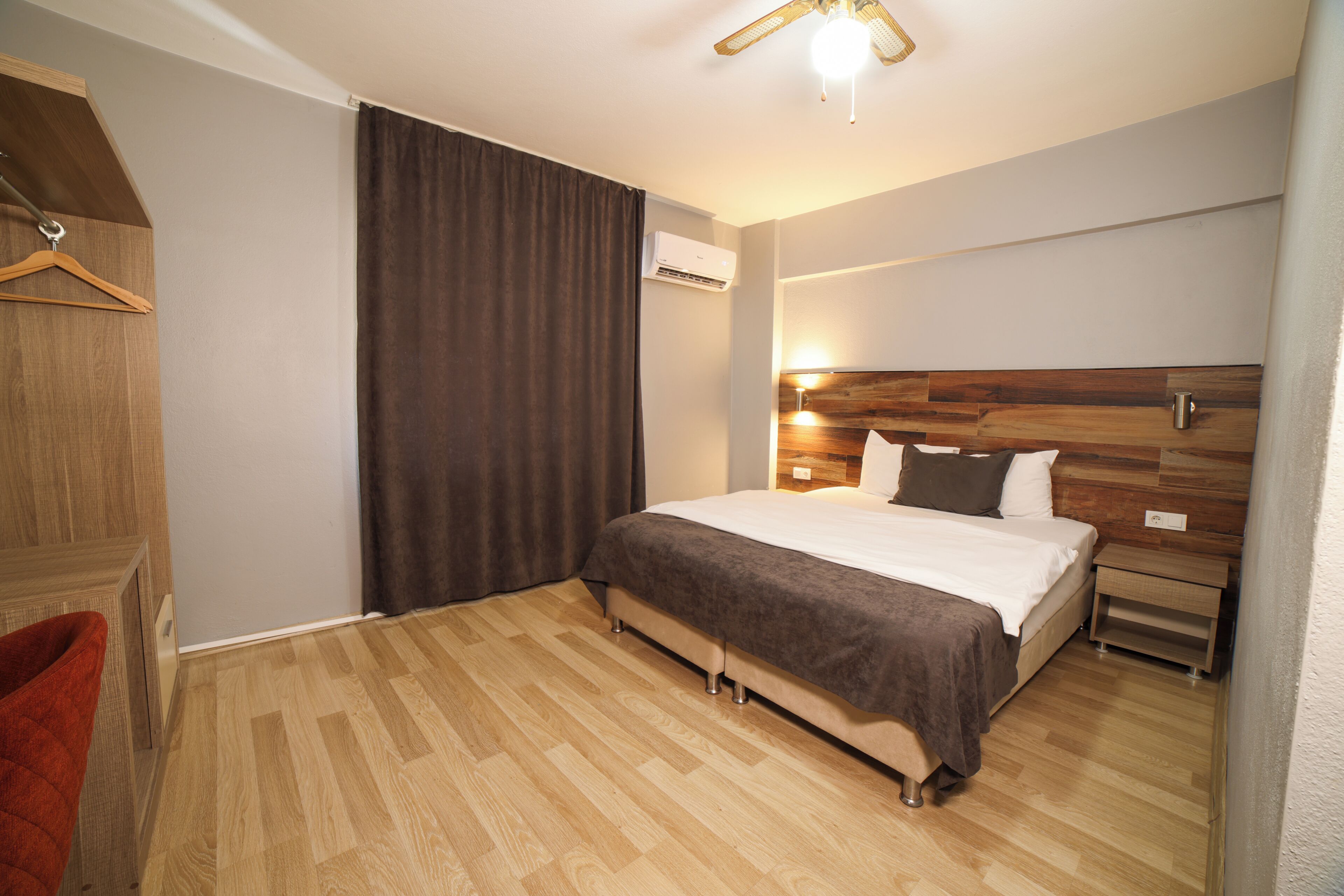 Un dormitorio con cabecera de madera, cama con manta marrón, suelo de madera, una silla colgante y un equipo de aire acondicionado.