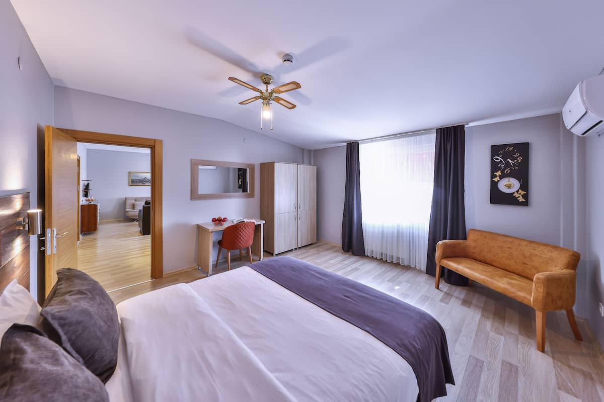 A modern hotel room with a large bed, a desk, a chair, a sofa, a ceiling fan, and a window with curtains.