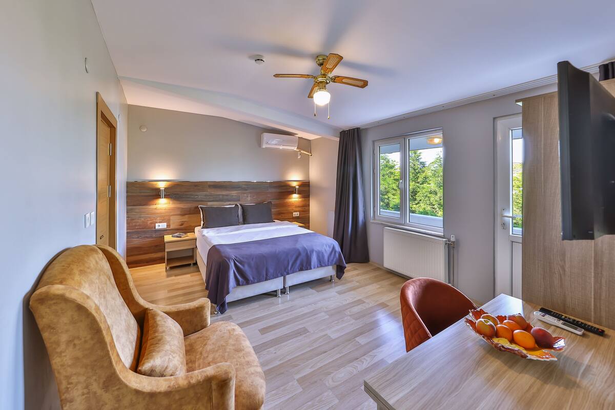 A modern hotel room with a bed, a desk, a chair, a ceiling fan, and a window with a view of greenery.