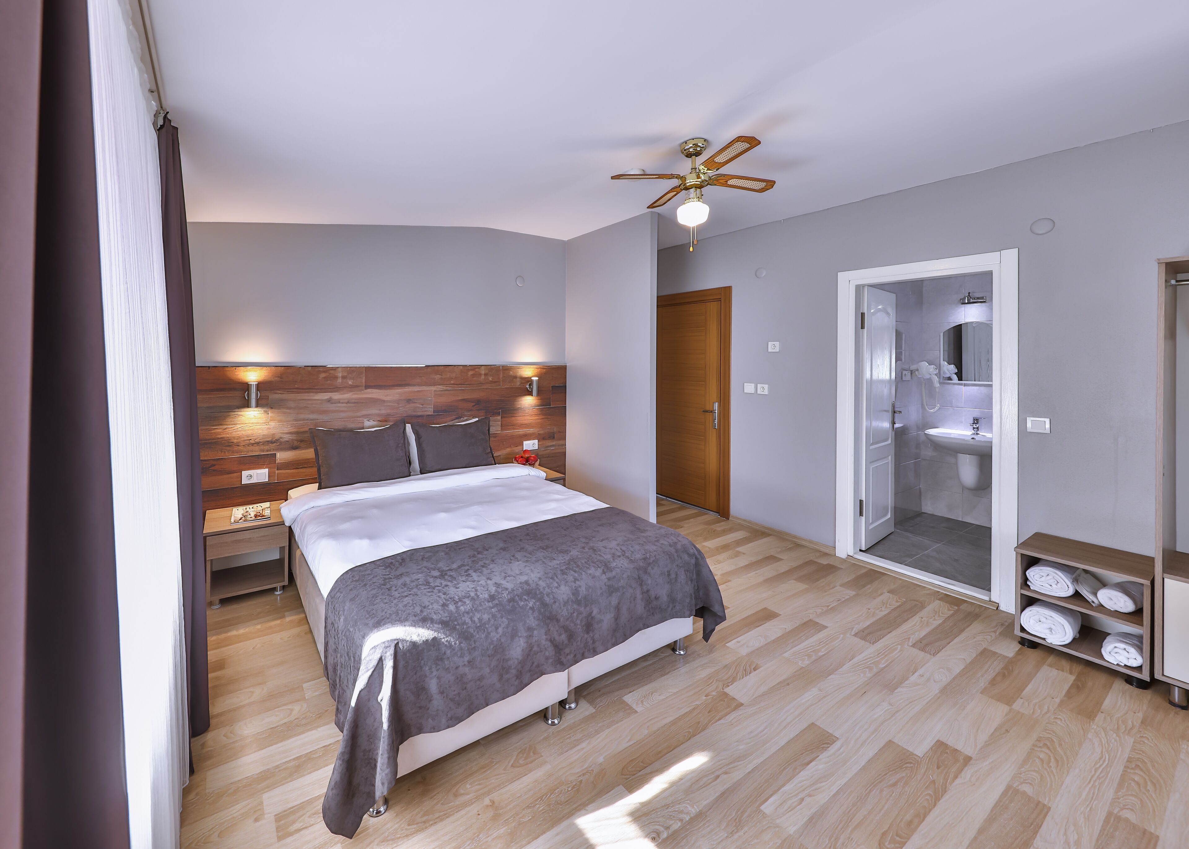 Un dormitorio moderno con cabecera de madera, cama con manta gris, ventilador de techo y vestidor con espejo.