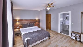 Un dormitorio moderno con cabecera de madera, cama con manta gris, ventilador de techo y vestidor con espejo.