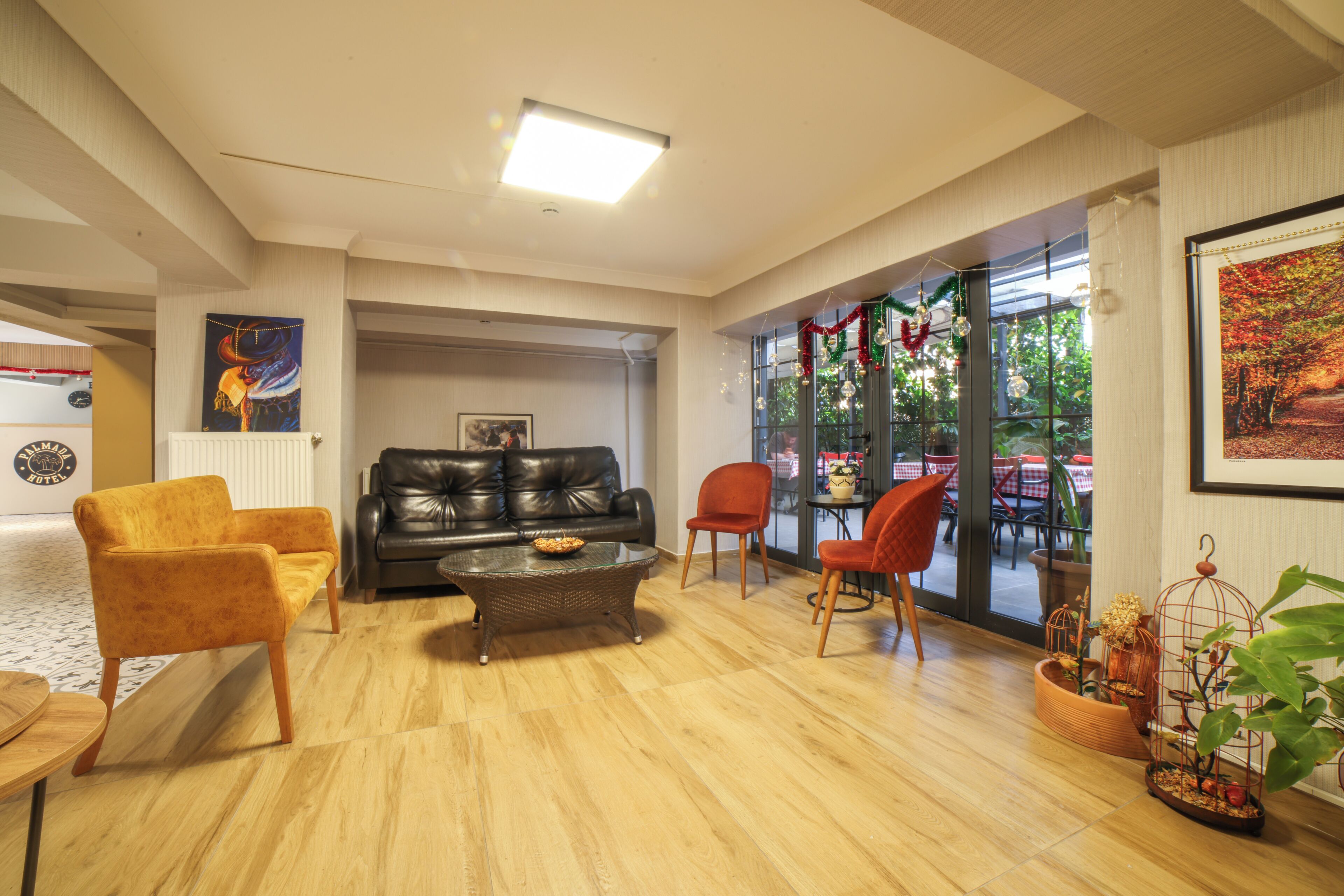 A spacious living room with a black leather sofa, a yellow armchair, and a glass coffee table. There are red chairs and a large window with a view of outdoor tables.