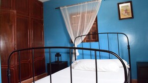 Un dormitorio con cama, mesita de noche, armario de madera, ventana con cortina translúcida y un cuadro en la pared.