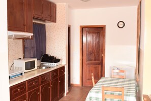 Een kleine keuken met houten kastjes, een magnetron en een spoelbak. Er is een deur naar een andere kamer en een klok aan de muur.