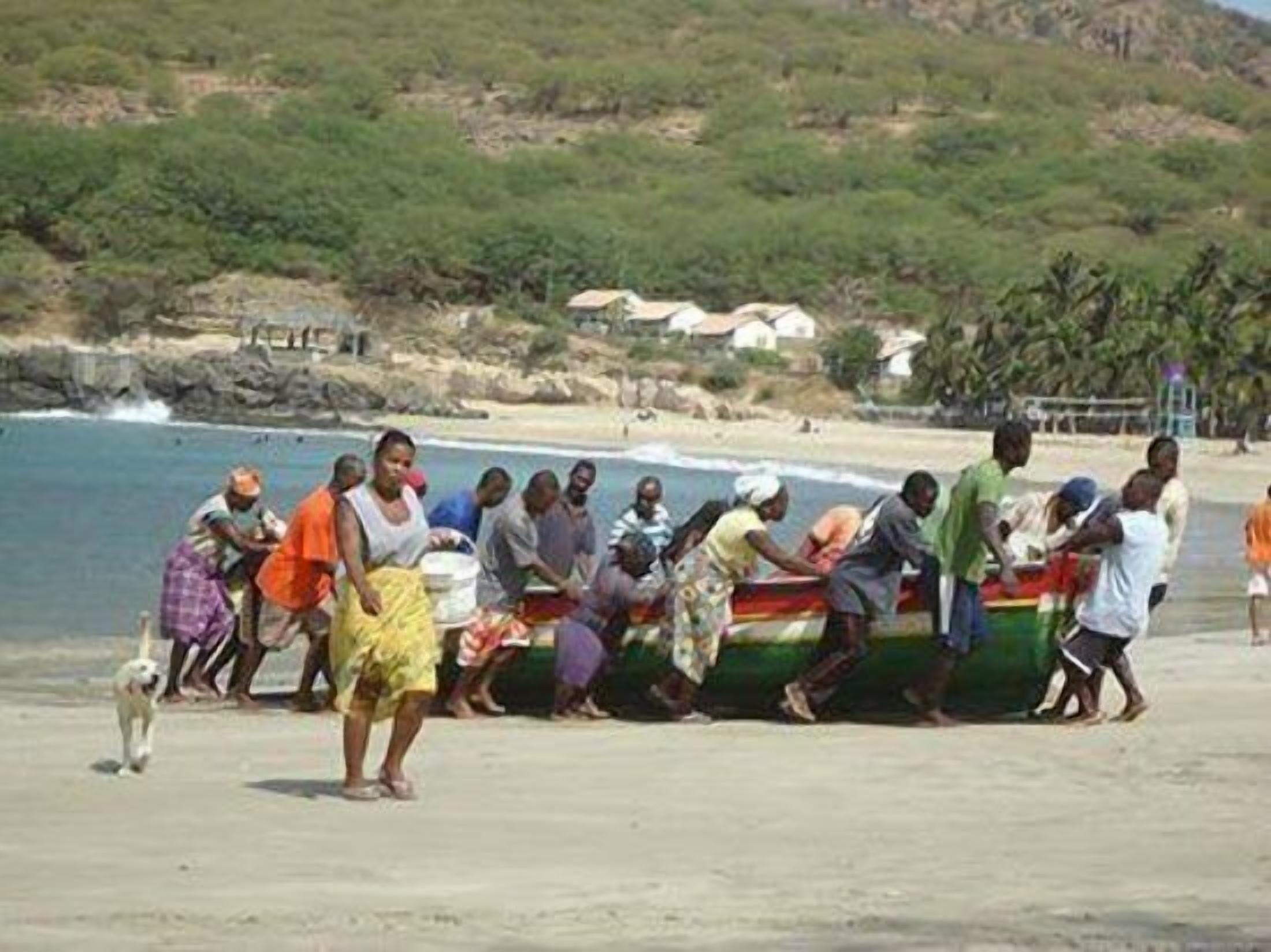A group of people gathered around a boat on a beach with a rocky shoreline and some greenery in the background.