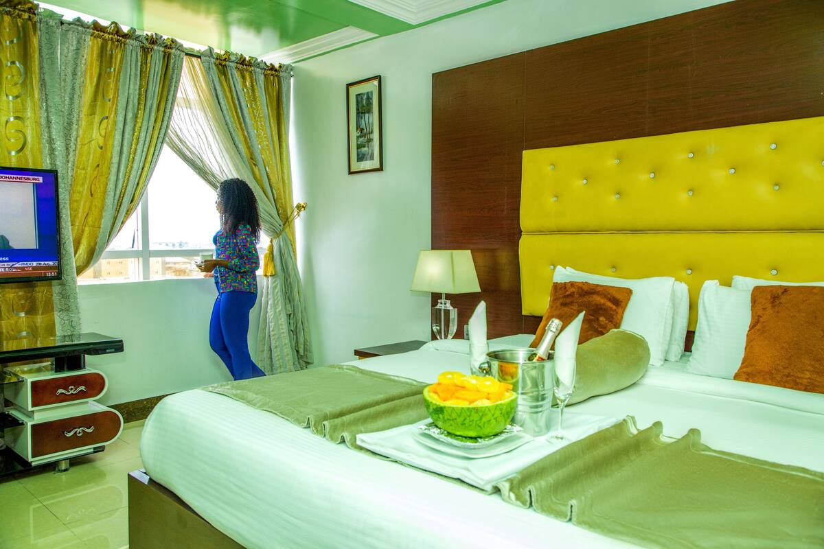 Une chambre d’hôtel avec un lit, une télévision, une fenêtre avec des rideaux, une table de chevet avec une lampe et un bol de fruits sur le lit.