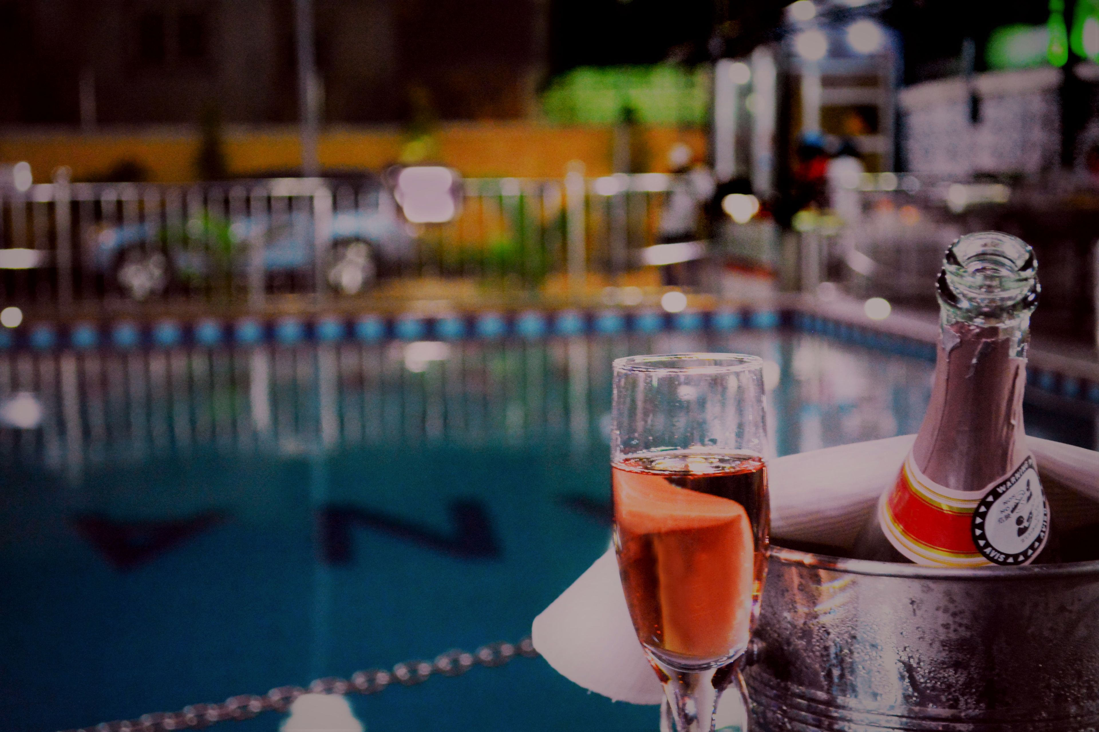 A glass of red wine and a bottle in a metal bucket by a pool.