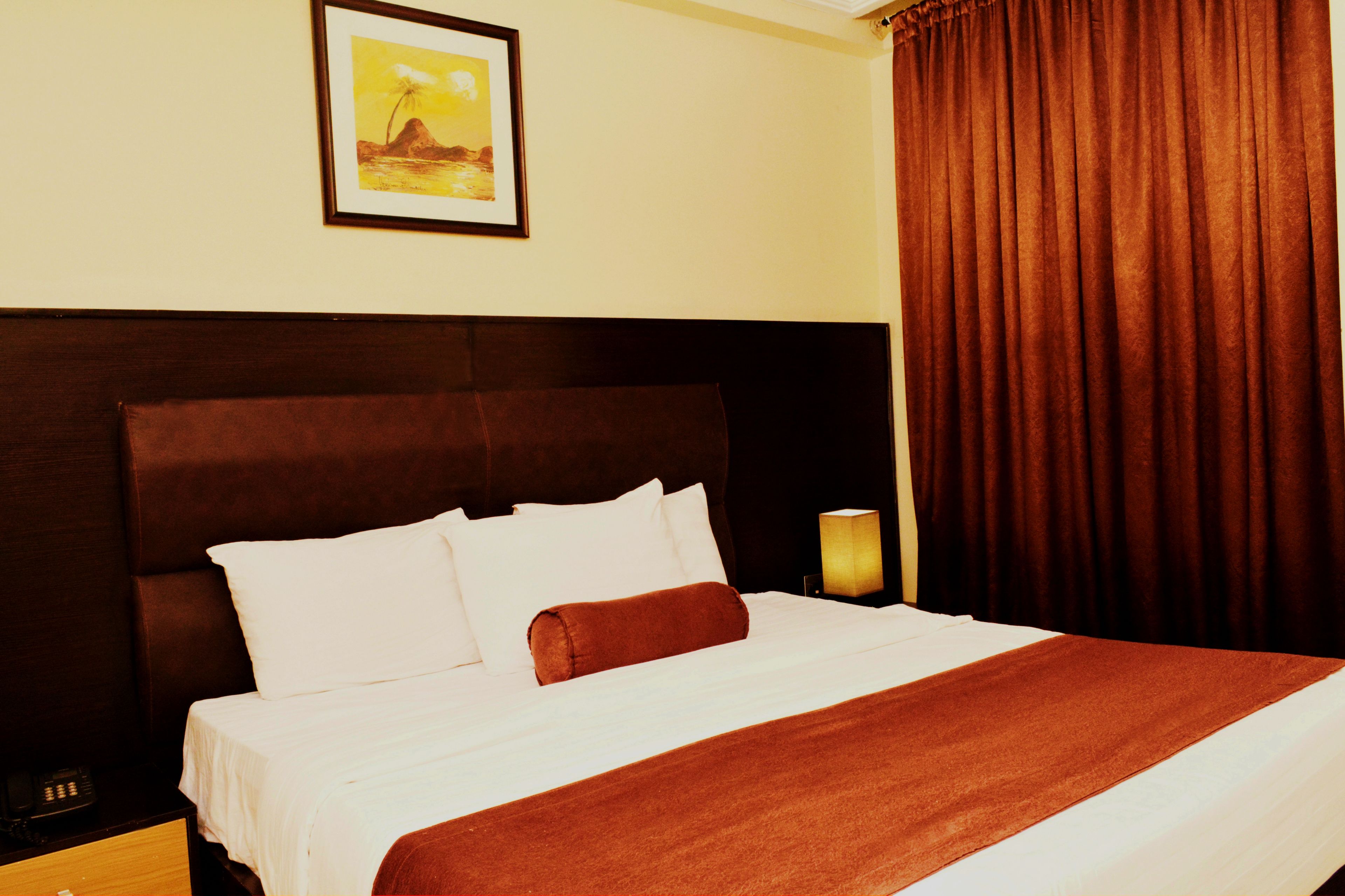A hotel room with a large bed, white bedding, a brown headboard, a framed picture on the wall, and a window with brown curtains.