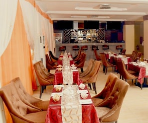 Um restaurante com mesas compridas, cadeiras estofadas em tons de marrom e um bar ao fundo.
