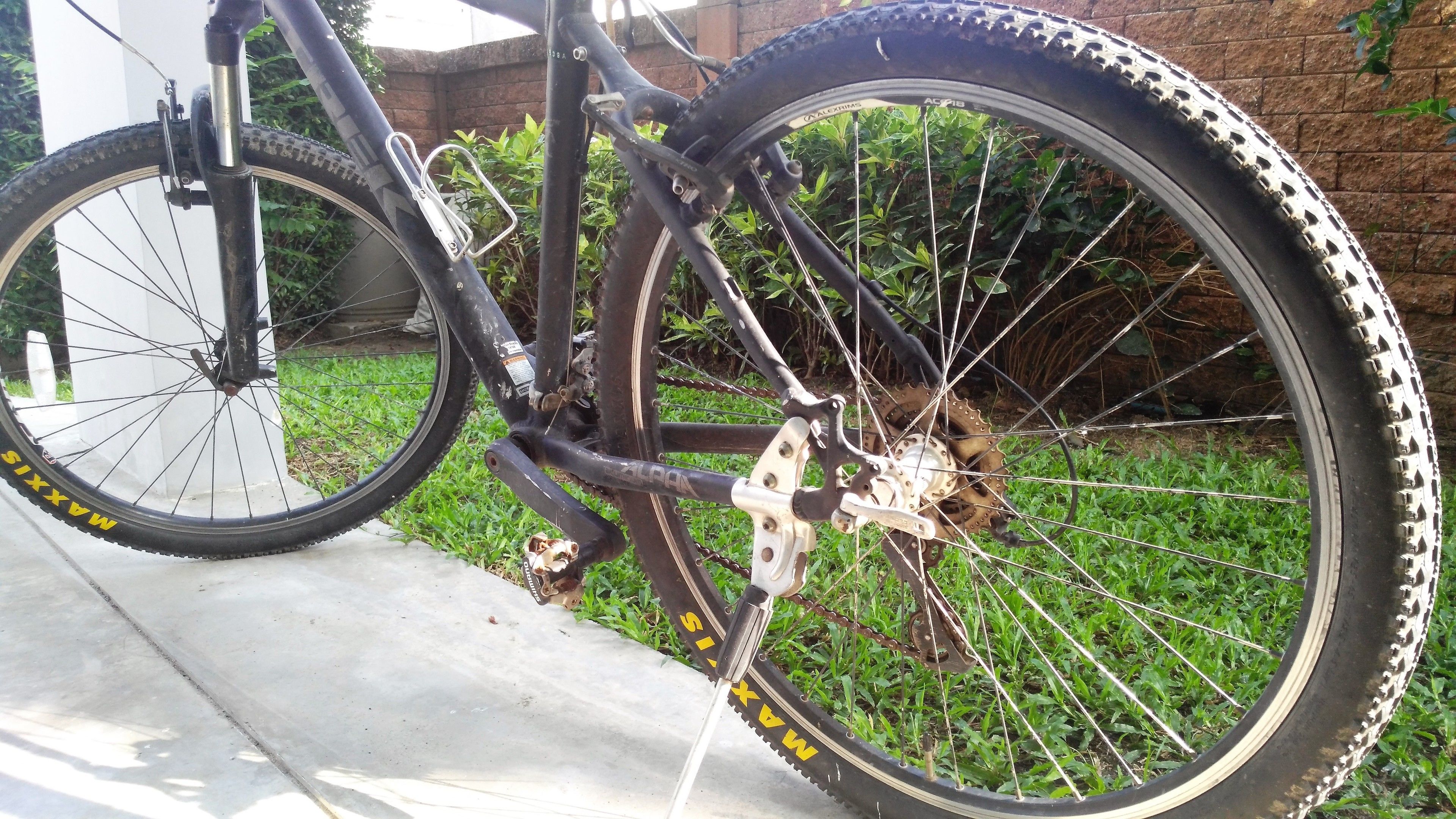 A bicycle with a rear derailleur and a cassette, parked on a concrete surface with grass and a brick wall in the background.