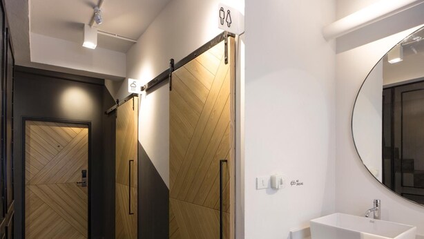 A modern bathroom with a wooden door, a white sink, and a mirror. - Bed One Block Hostel (Bangkok)