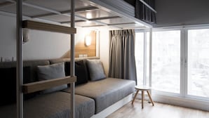 A modern, compact room with a sofa bed, a small wooden stool, and a large window offering a view of the city. - Bed One Block Hostel (Bangkok)