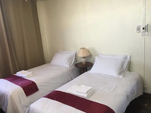 A hotel room with two single beds, each with a white comforter and a red throw blanket. There is a small bedside table with a lamp and a phone charger.