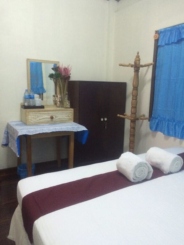 A bedroom with a bed, a dresser with a mirror, a wooden wardrobe, and a window with blue curtains. - Pancake House B&B (Prachuap Khiri Khan)