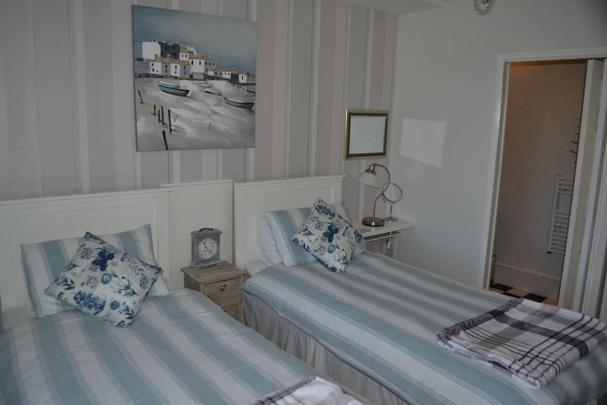 A bedroom with two beds, striped bedding, a painting of a coastal scene, a bedside table with a lamp, and a door leading to another room.