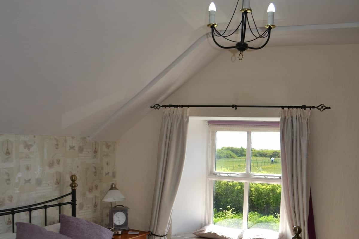 A bedroom with a bed, a nightstand, a lamp, a window with curtains, and a chandelier.