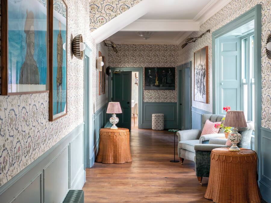 Un couloir avec un papier peint à motifs, des tables d’appoint en osier, un canapé et des œuvres d’art encadrées accrochées aux murs.