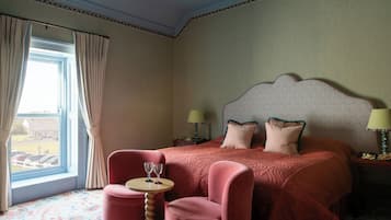Une chambre d’hôtel avec un grand lit, deux lampes de chevet, une petite table ronde avec deux verres, un fauteuil rose et une fenêtre avec des rideaux.