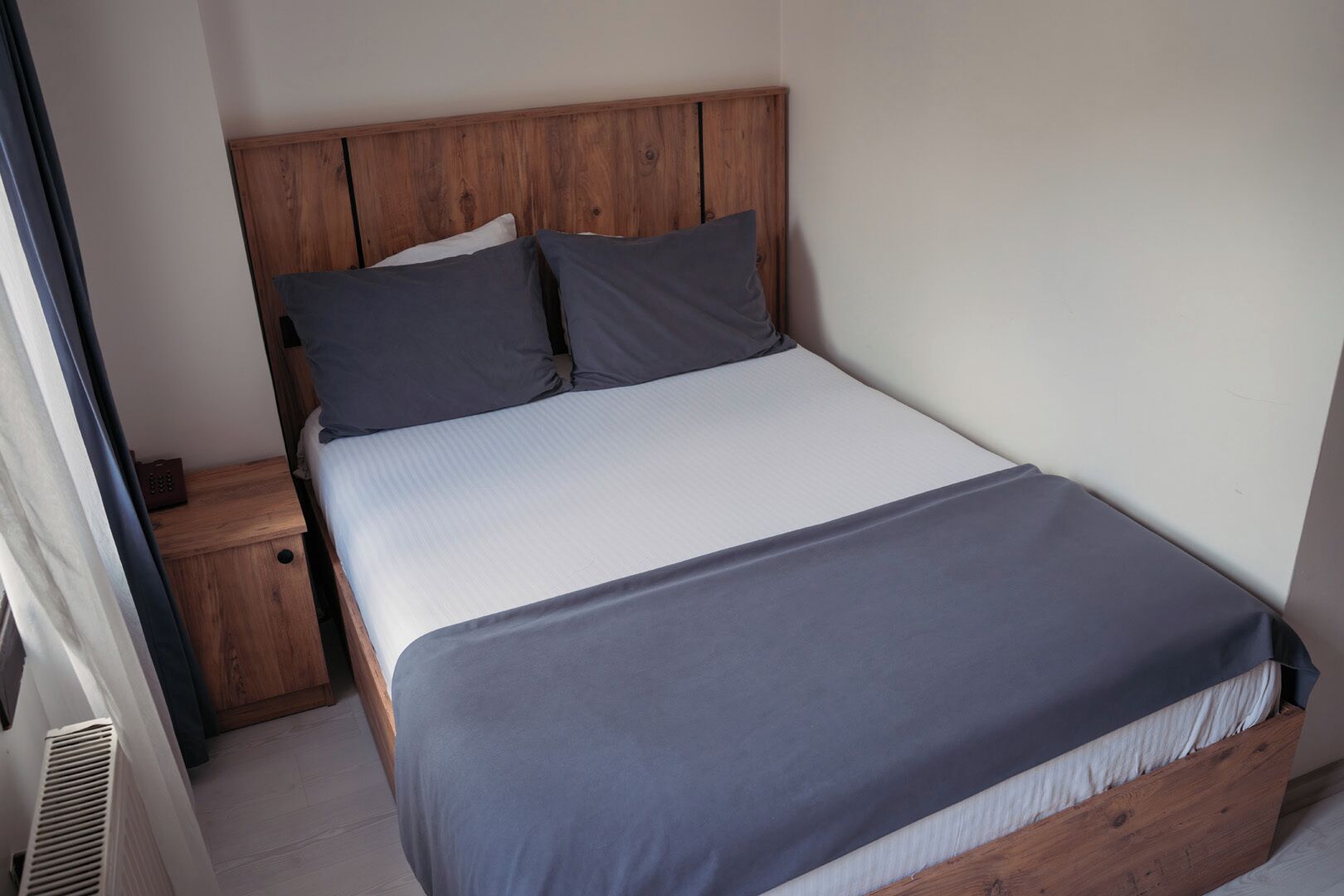 A single bed with a wooden headboard, a bedside table, and a grey blanket.