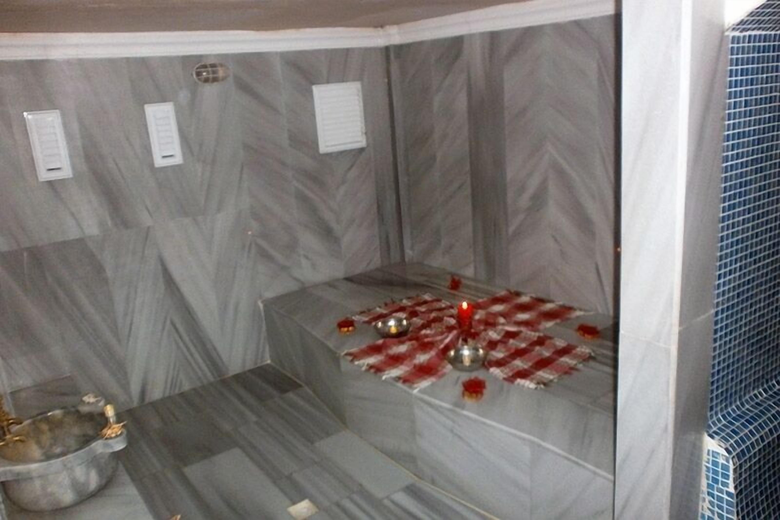 A small, enclosed room with marble walls and a tiled floor. There is a small table with a red candle and a metal bowl.