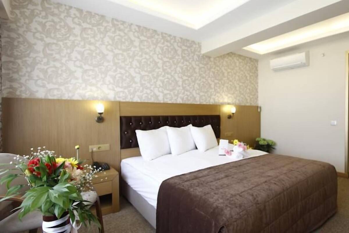 A hotel room with a large bed, a nightstand with a lamp, a potted plant, and a door to the right.