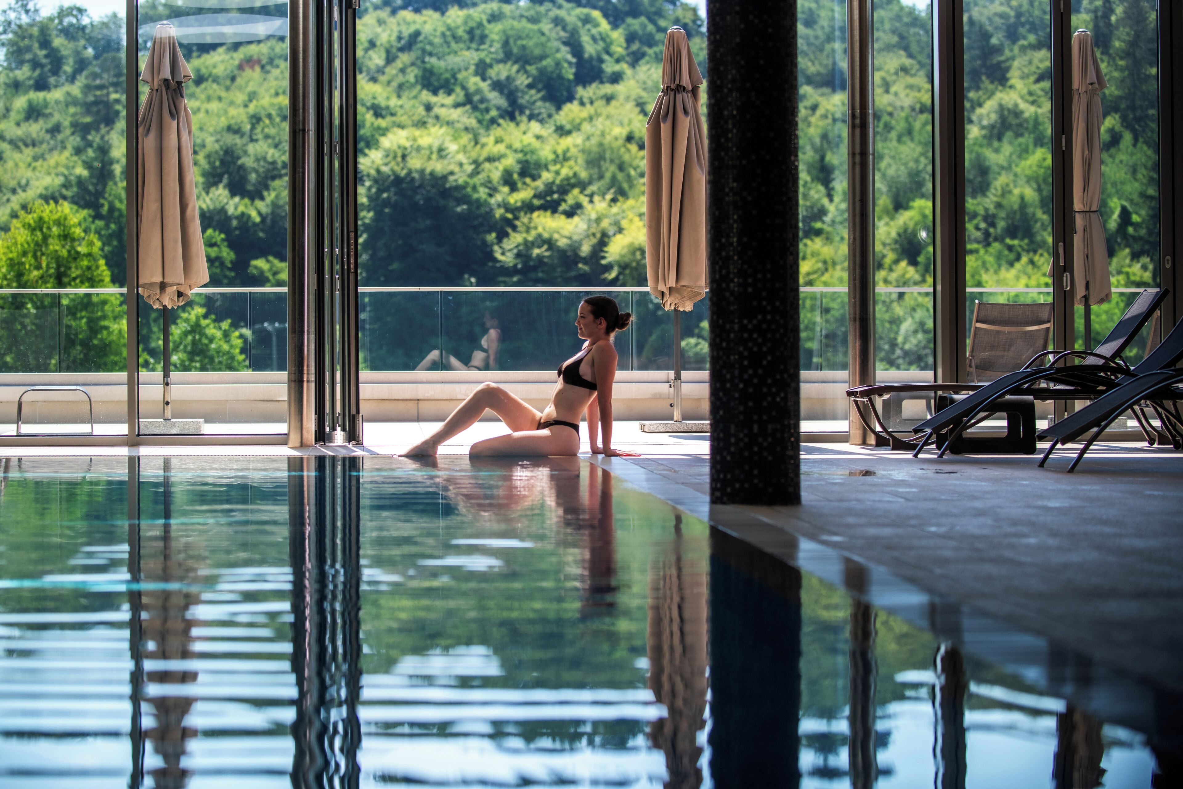 A pool area with a person sitting by the poolside, surrounded by lounge chairs and umbrellas.