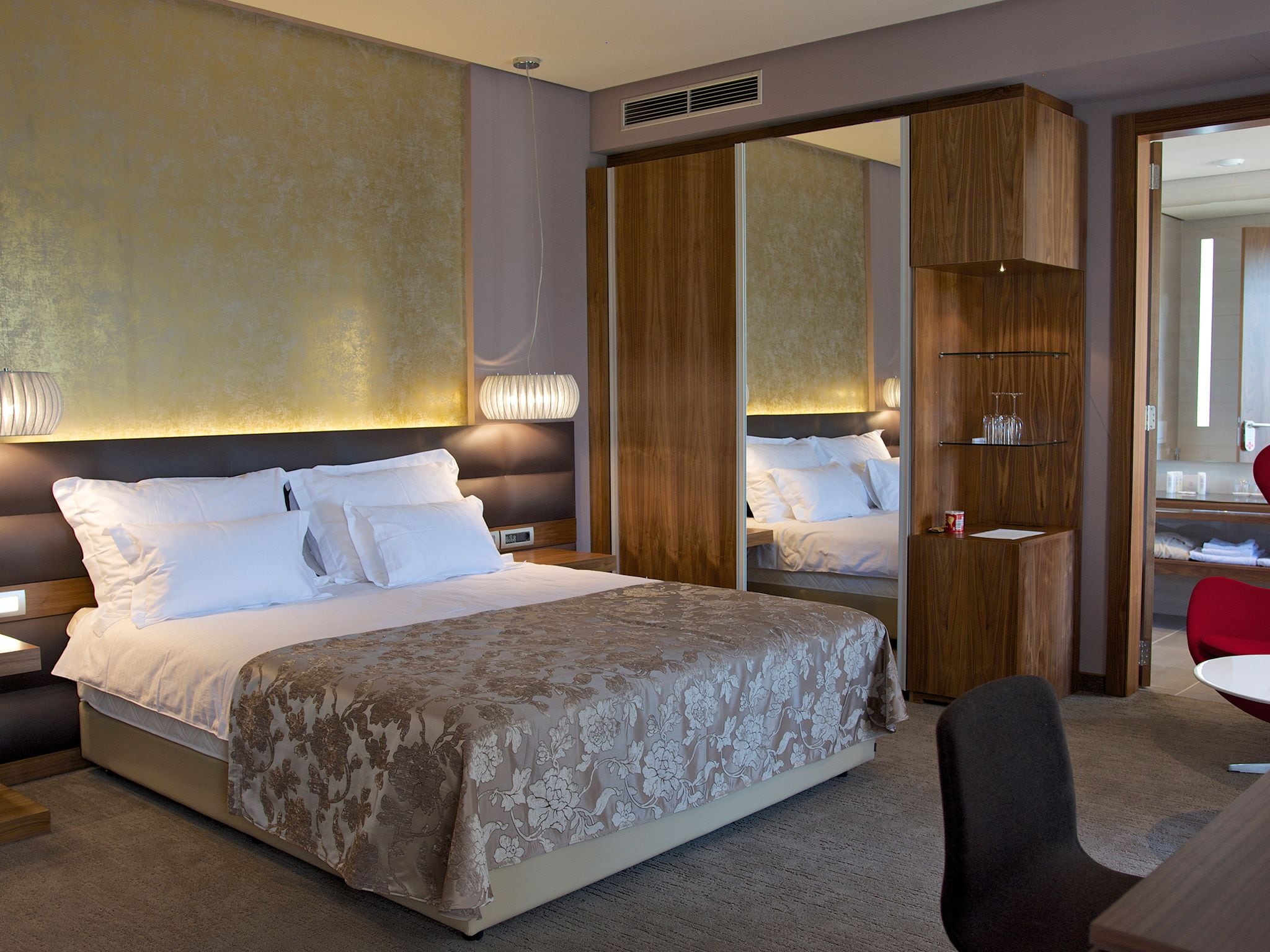 Quarto de hotel moderno com uma cama grande, guarda-roupa de madeira, espelho e uma pequena mesa com cadeira.