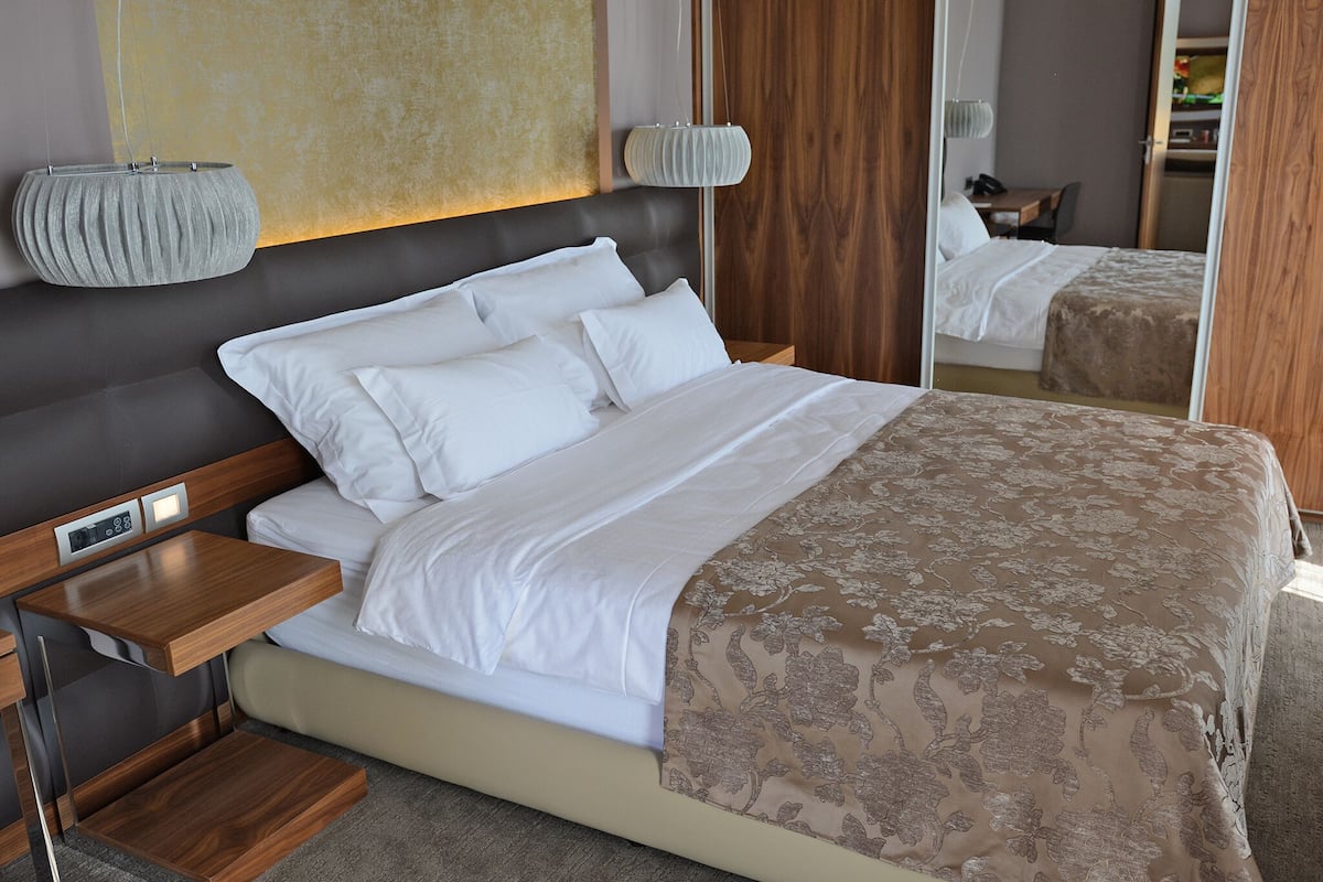 A modern hotel room with a large bed, bedside tables, a mirror, and a wooden headboard.