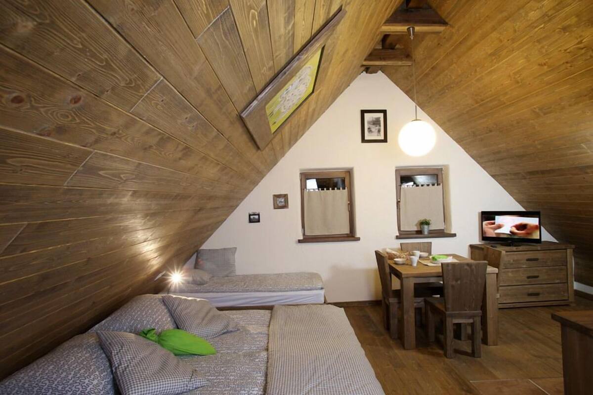 A cozy attic bedroom with a sloped ceiling, wooden walls, a bed with patterned bedding, a desk with a chair, and a television.