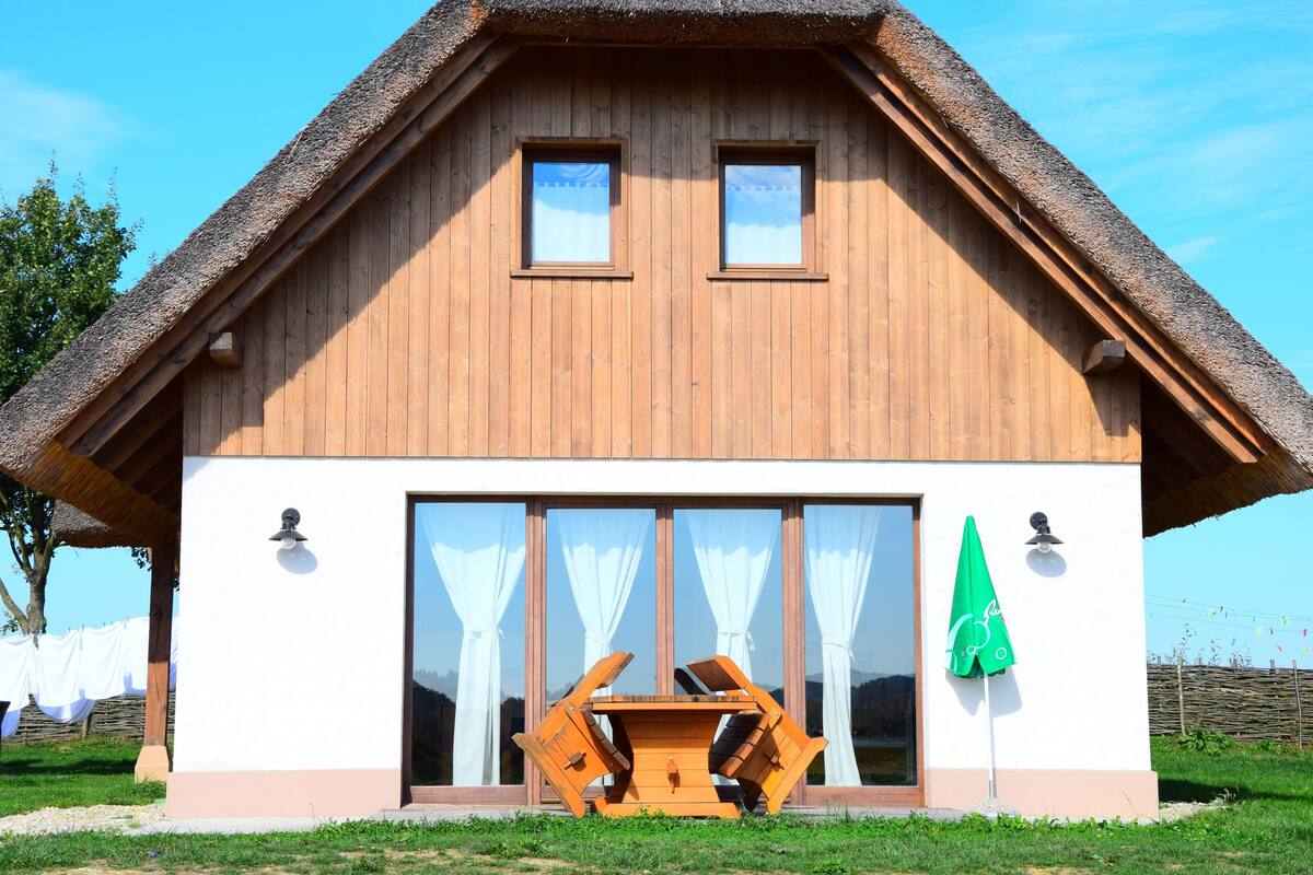 A wooden house with a thatched roof, large windows, and a small outdoor seating area.