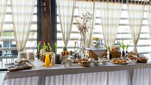 Mesa de desayuno buffet con diversos alimentos, como frutas, pasteles y bebidas, montada en una sala con ventanas y cortinas grandes.