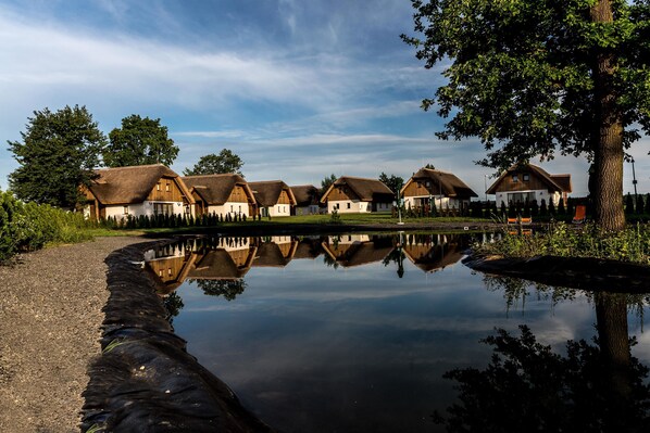 A row of traditional houses with thatched roofs reflected in a calm body of water. - Panonska Vas Villas and Studios (Moravske Toplice)