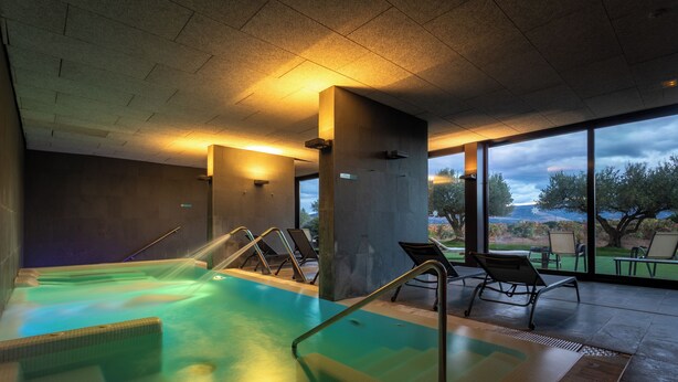 Un área de piscina interior moderna con paredes de vidrio transparente, vistas al exterior y algunas sillas de descanso.