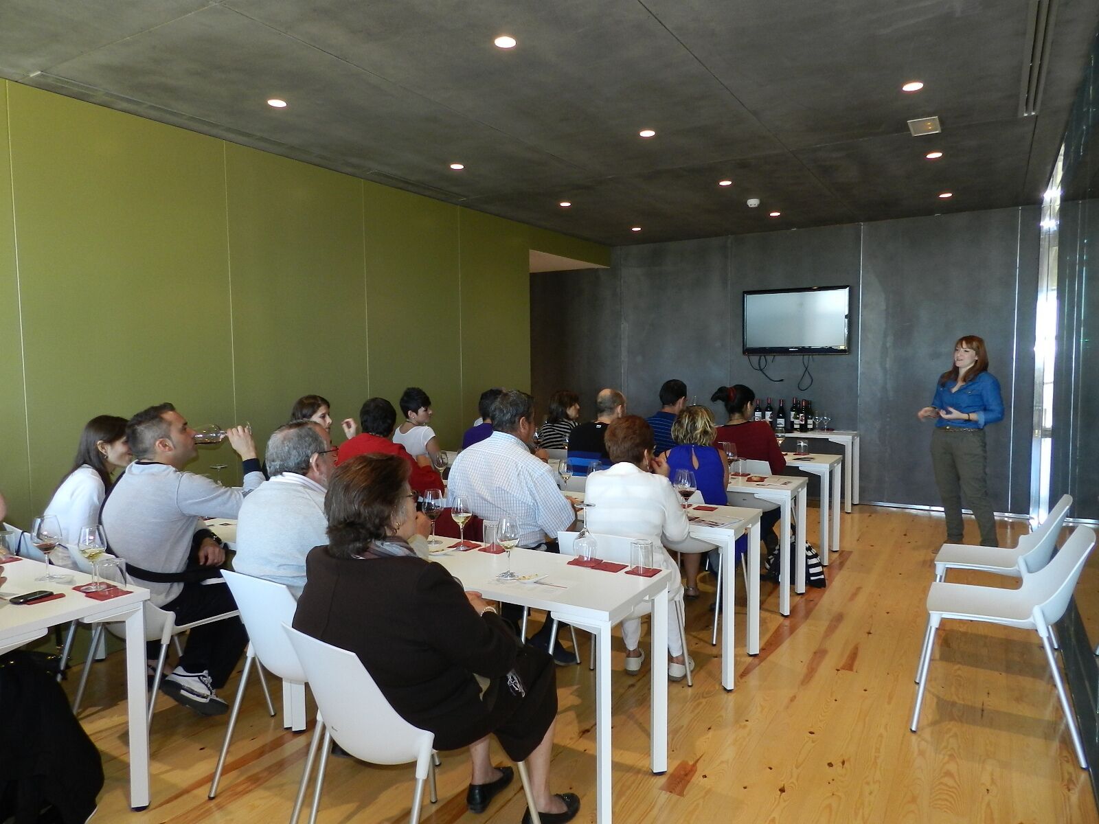 A conference room with attendees seated at white tables, a speaker at the front, and a large screen on the wall.