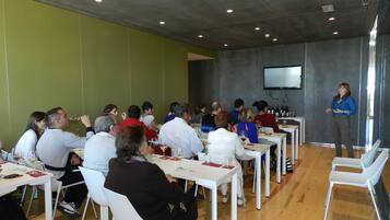 A conference room with attendees seated at white tables, a speaker at the front, and a large screen on the wall.