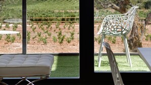 Un balcón con una silla blanca y un banco acolchado, con vistas a un viñedo y montañas lejanas.