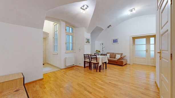 A spacious room with wooden flooring, a dining area with chairs and a table, a sofa, and a large window.